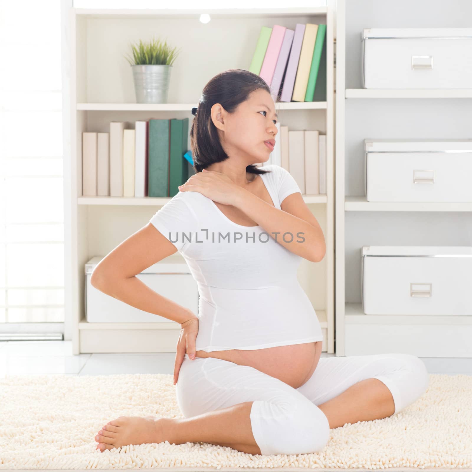 Shoulder or neck pain. Eight months pregnant Asian woman holding her back and shoulder while sitting on a floor at home.