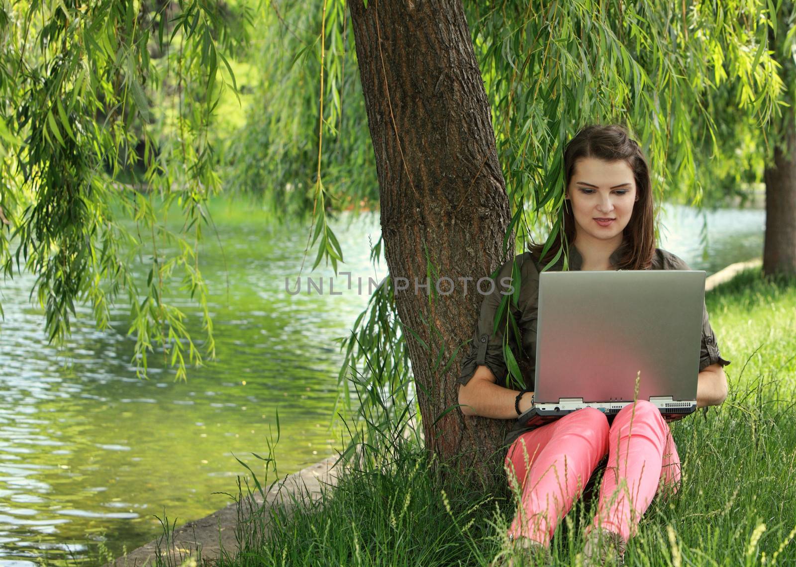 Young preety woman studying outside in a park under a willow tree, near a pond.