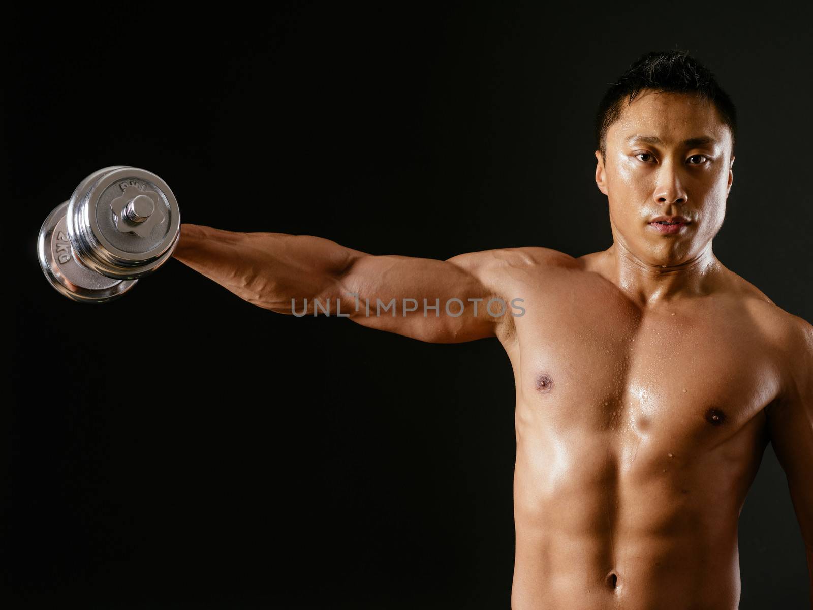 Photo of an Asian male exercising with dumbbells and doing shoulder flys or lateral flys over dark background.