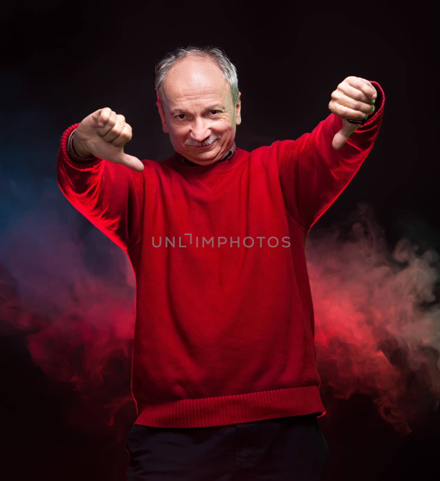 An elderly man in a red sweater shows thumbs down on a dark background