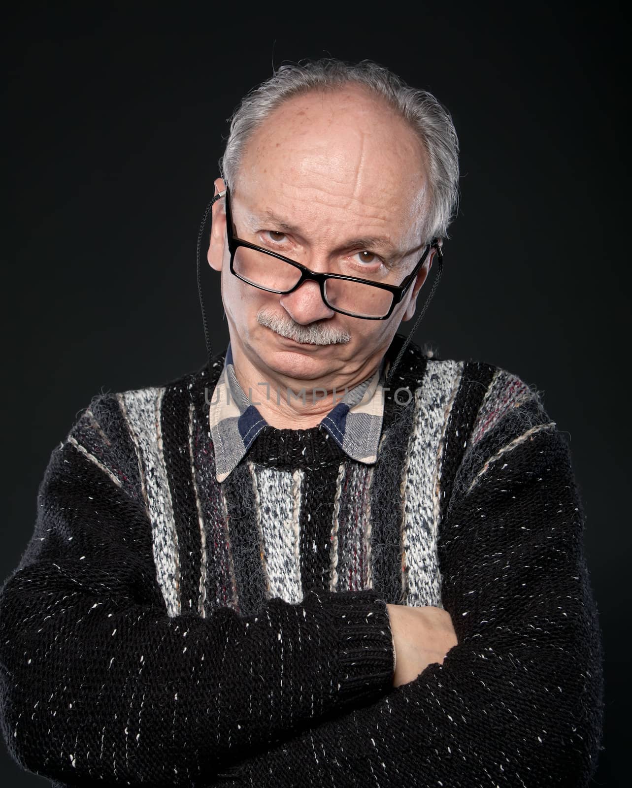 An elderly man with glasses and crossed arms looks skeptically on a dark background