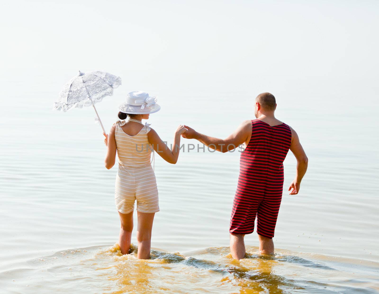 A man and a woman in vintage striped bathing suits go into the sea holding hands