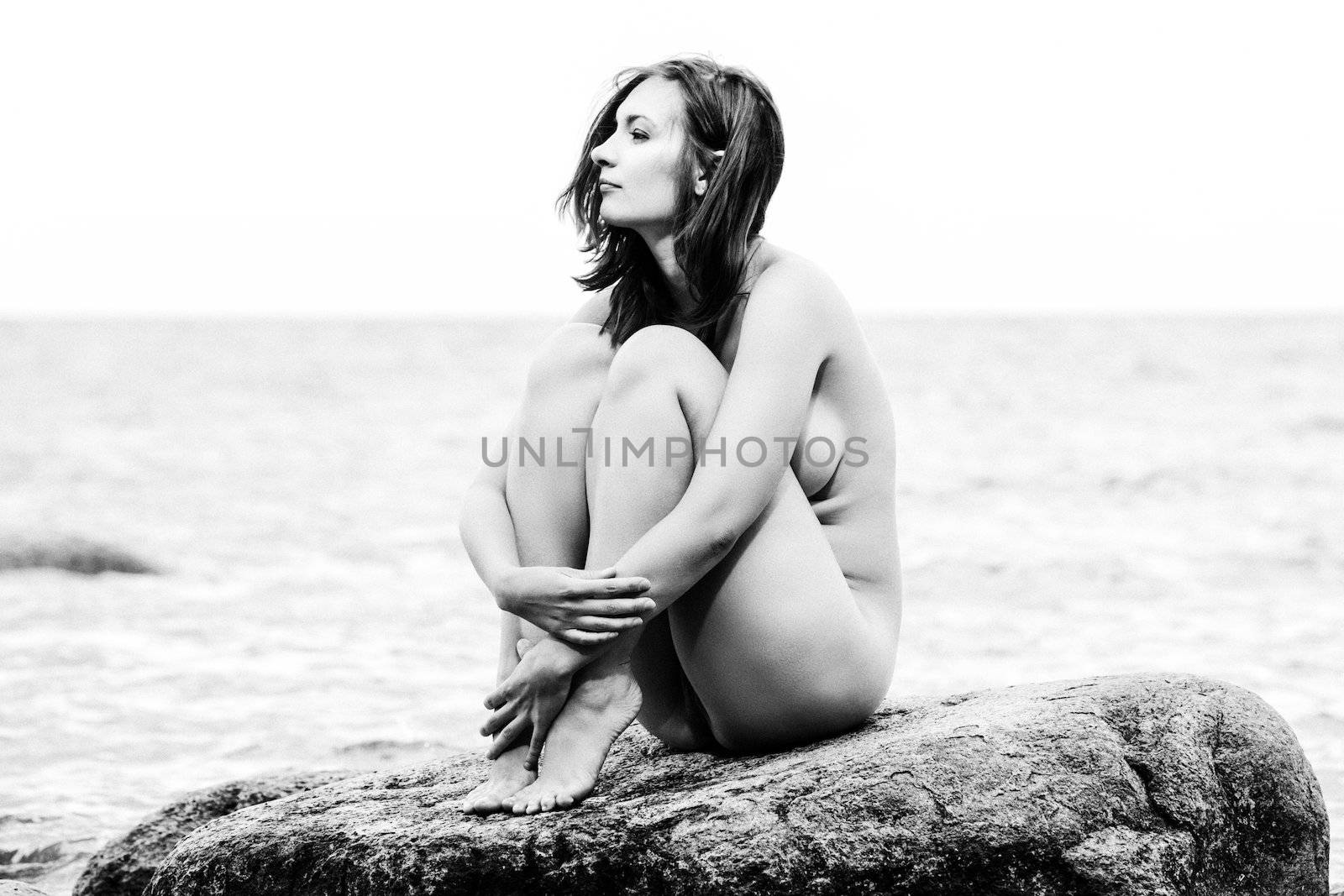 Young nude woman sitting on stone against the sea background. Old photo stylization. Film grain added