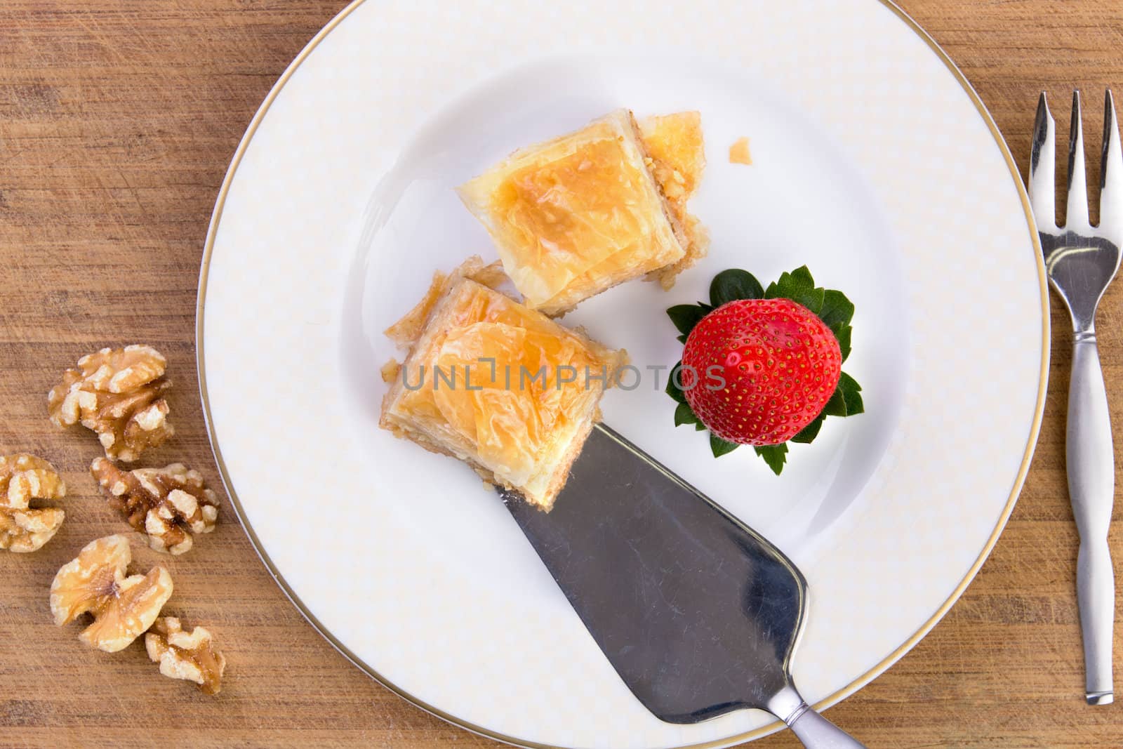 Couple pieces of home made baklava getting ready to be served with stainless-steel spatula on old bamboo cutting board along with walnuts and strawberry