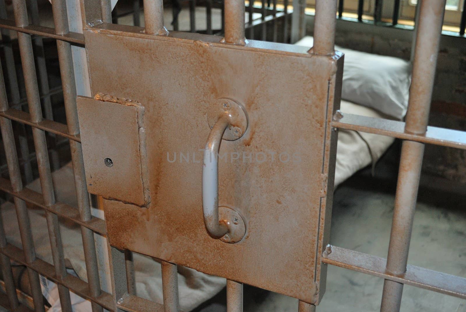 A view through a jail cell door bars revealing a small cot