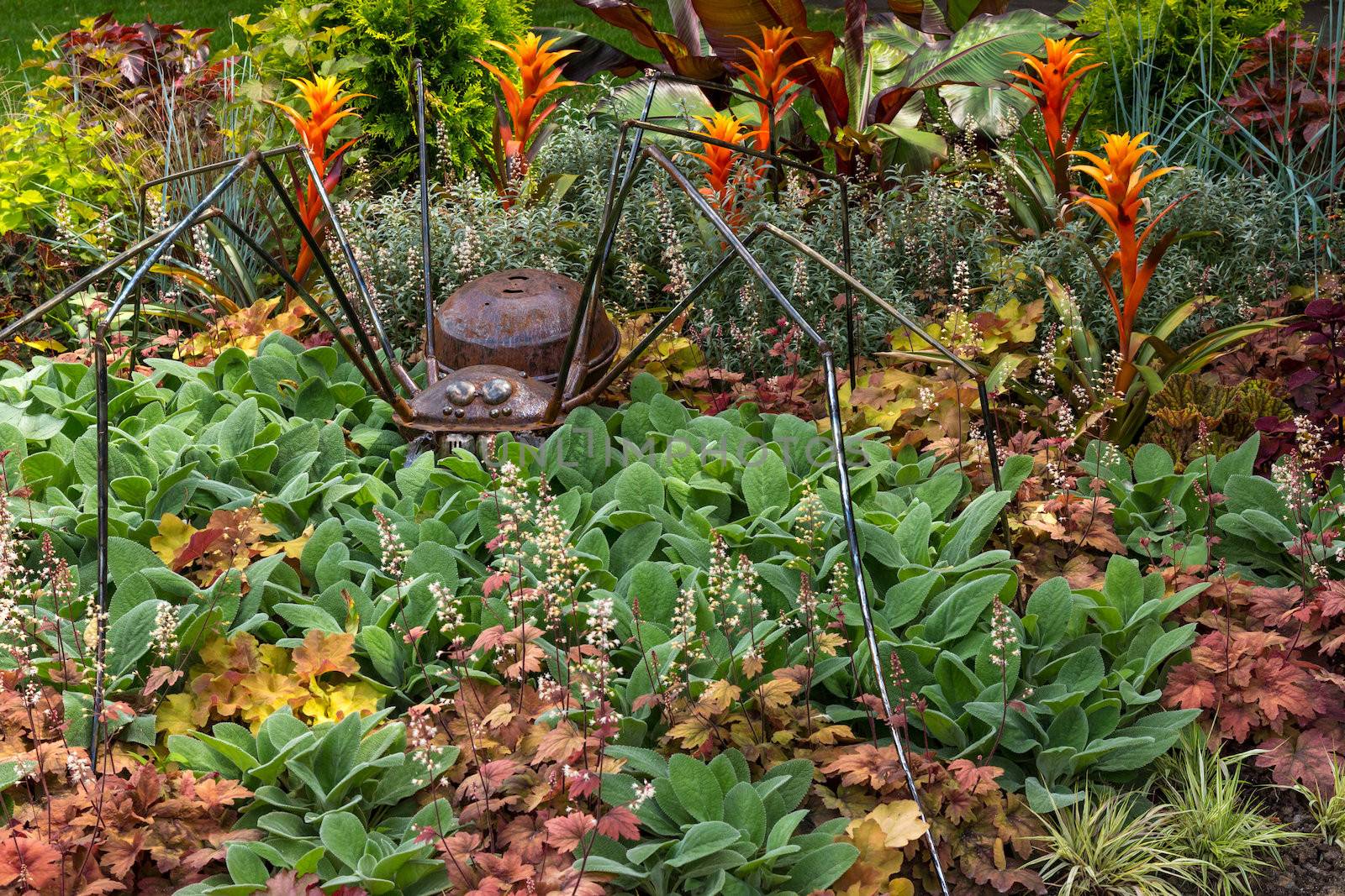 Rusty metal spider decorating a garden with colorful variety of plants.