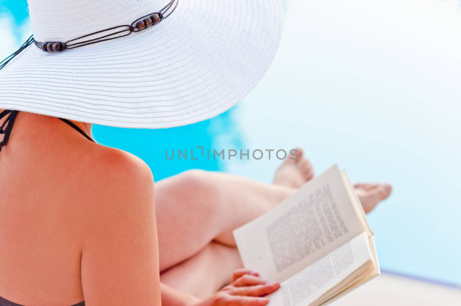 Girl in a hat reading a book near the pool, sitting in a lounge chair