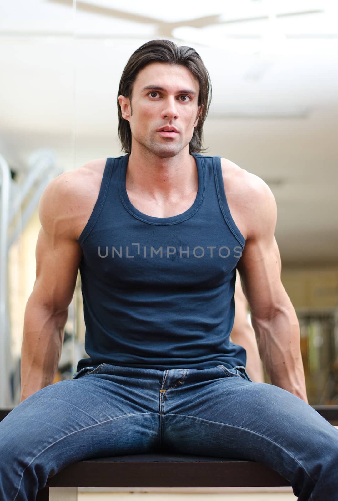 Attractive, muscular man sitting on desk indoors, wearing jeans and tanktop