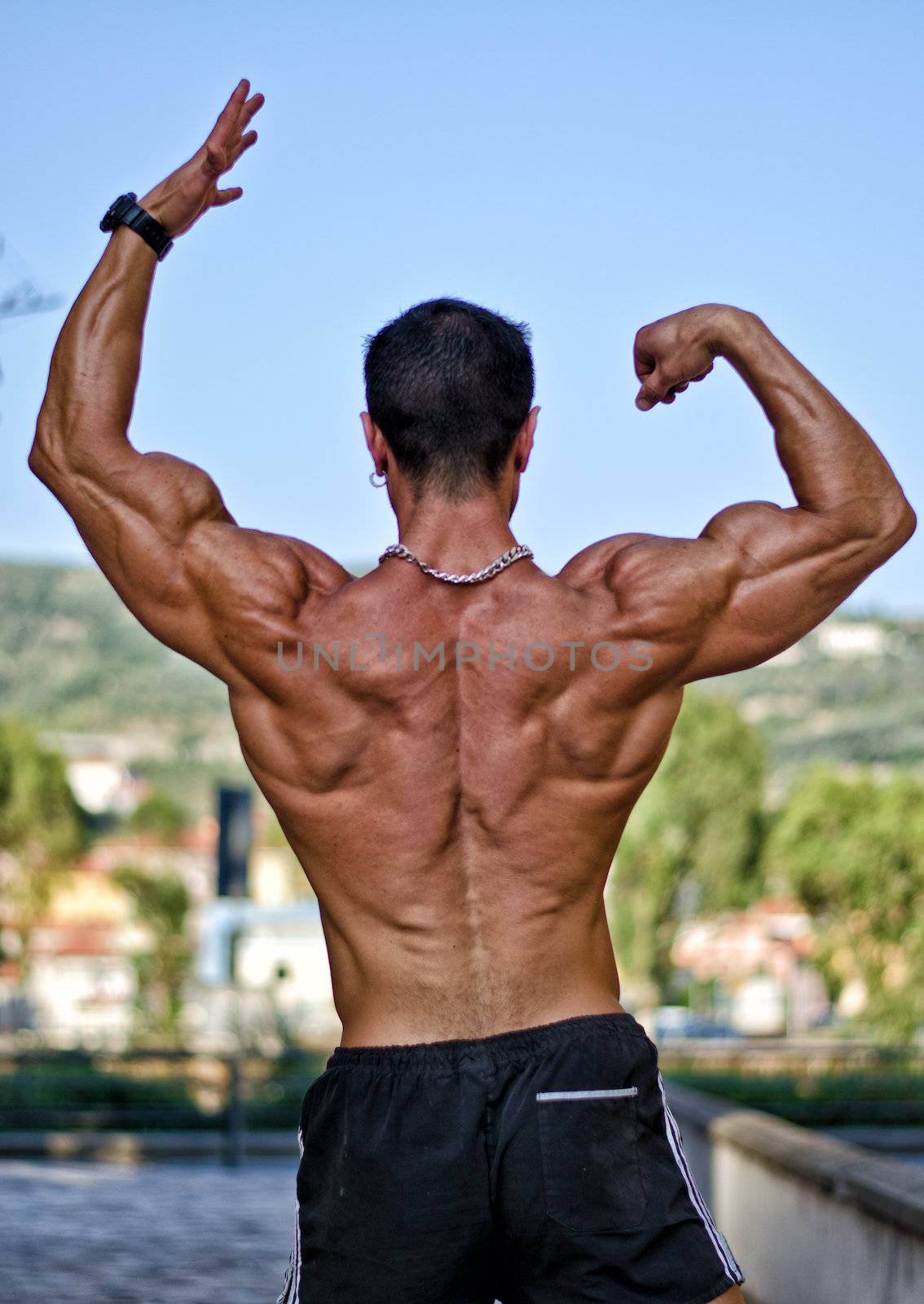 Back of muscular bodybuilder in classic pose, outdoors wearing black trunks