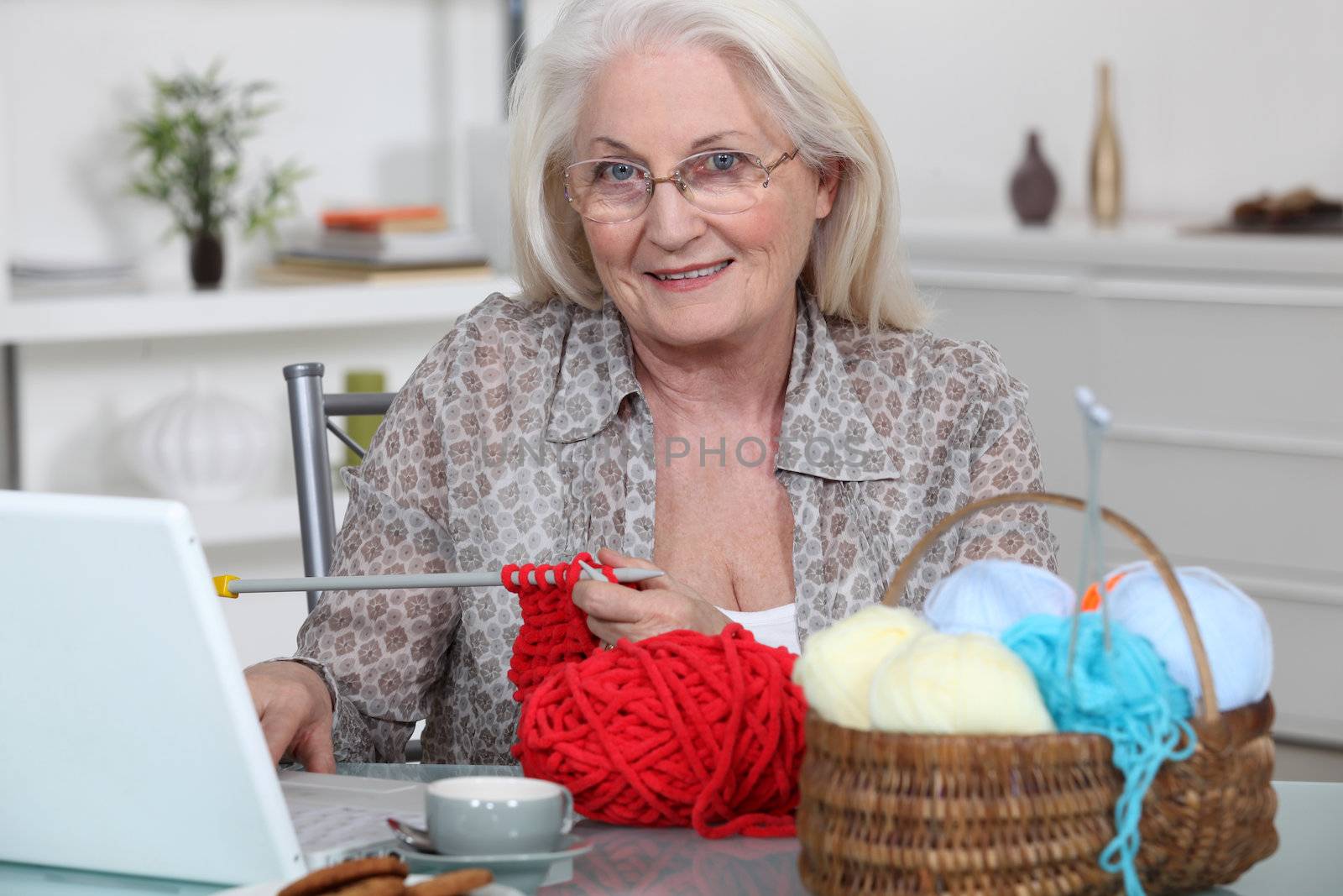Senior woman knitting in front of her laptop computer by phovoir