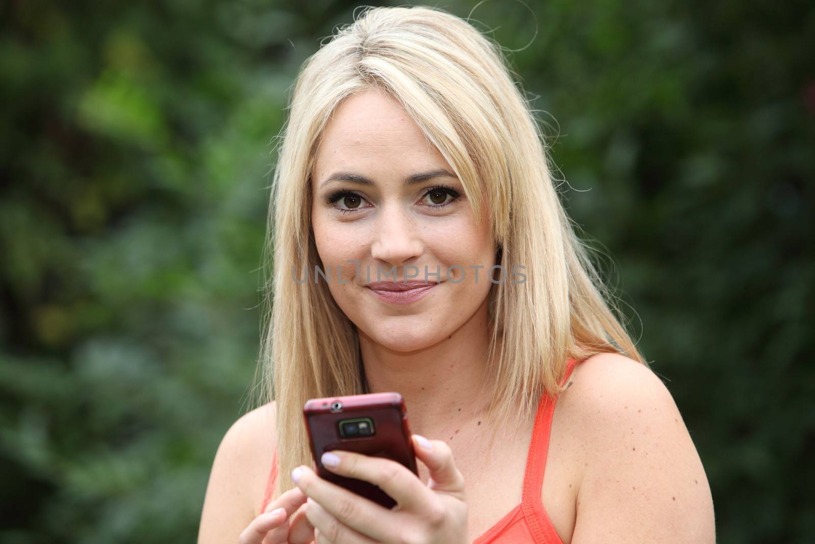 Smiling blond woman in a summer top standing in a green garden with a mobile phone in her hands looking at the camera