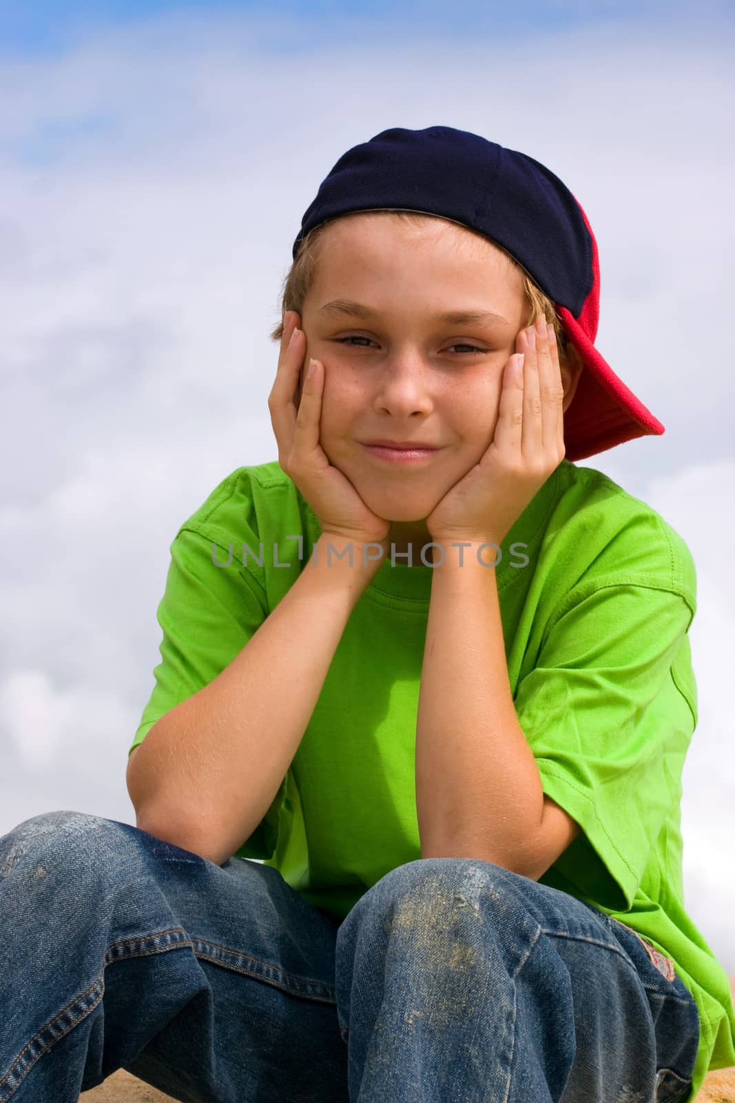 Smiling boy head in hands wearing jeans, t-shirt and baseball cap sitting outside.