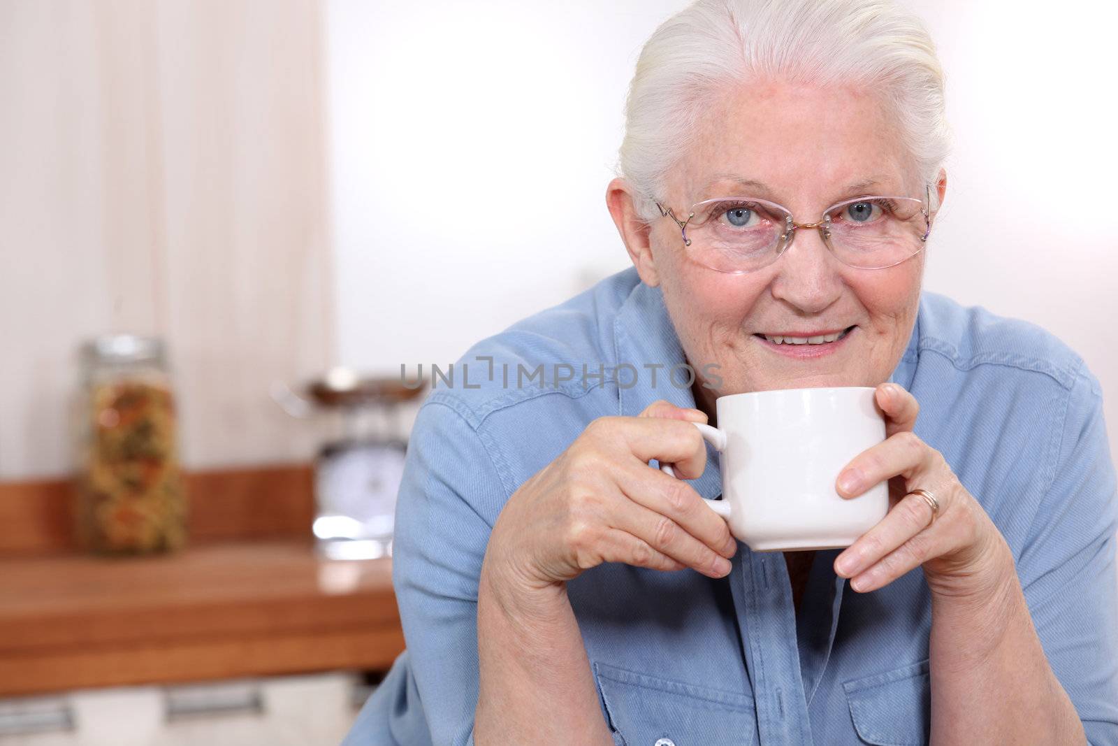 Elderly lady enjoying cup of tea in her kitchen by phovoir