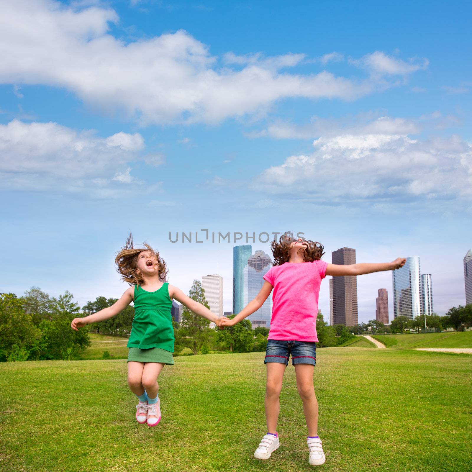Two sister girls friends jumping happy holding hand in urban modern skyline on park lawn