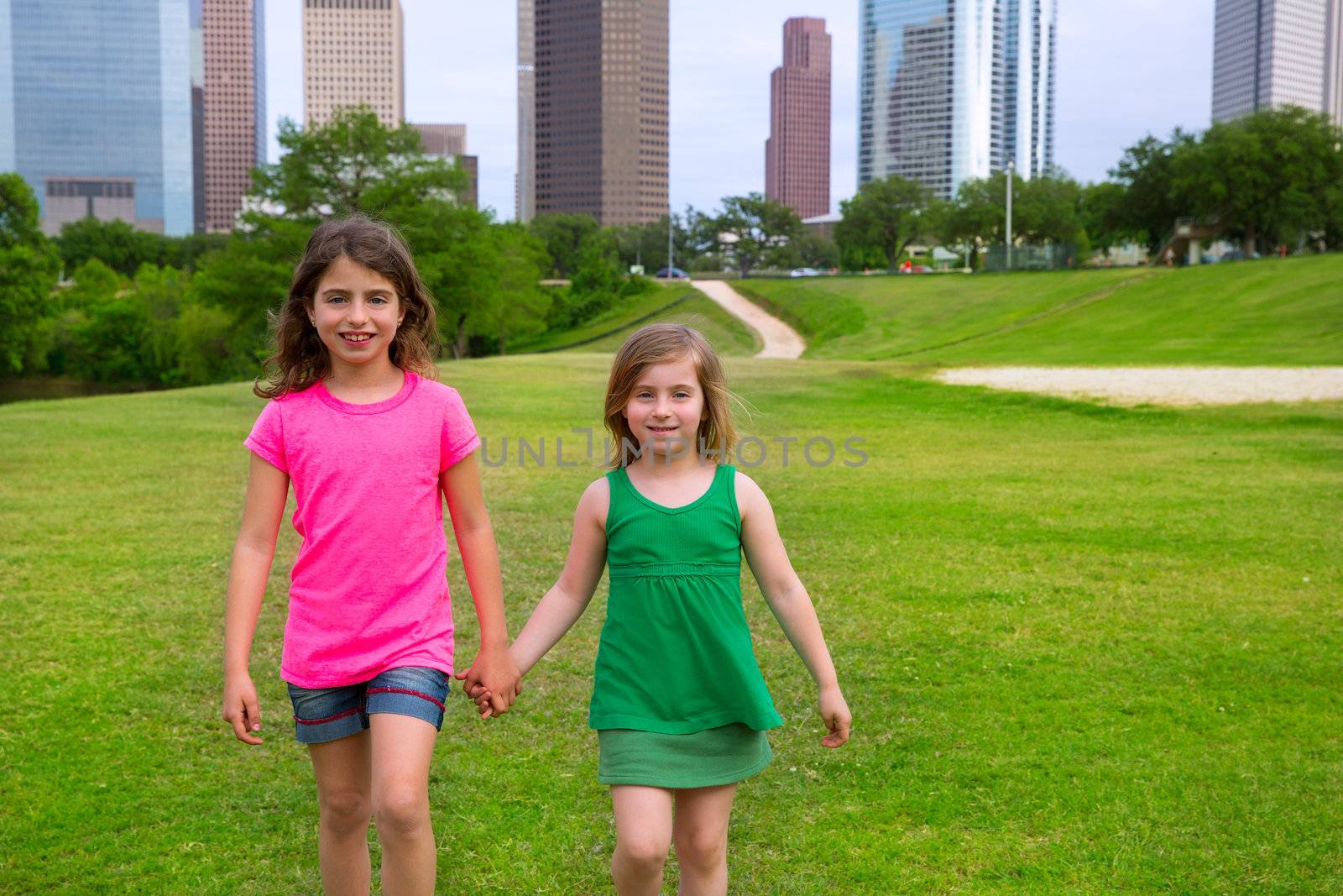 Two sister girls friends walking holding hand in urban modern skyline on grass lawn