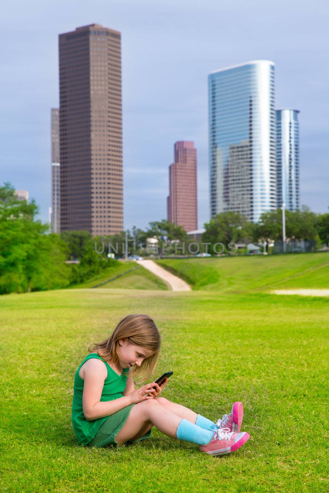 Blond kid girl playing with smartphone sitting on park lawn in city skyline background