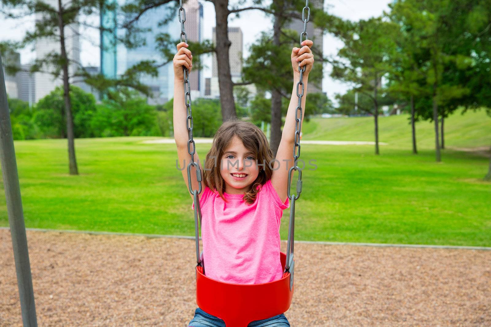 Brunette kid girl playing with swing on park with city skyline background