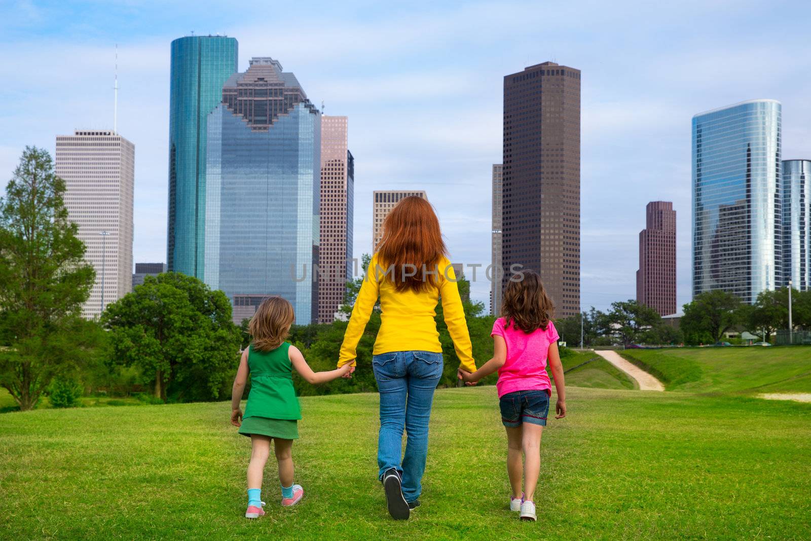 Mother and daughters walking holding hands on modern city skyline over park green lawn