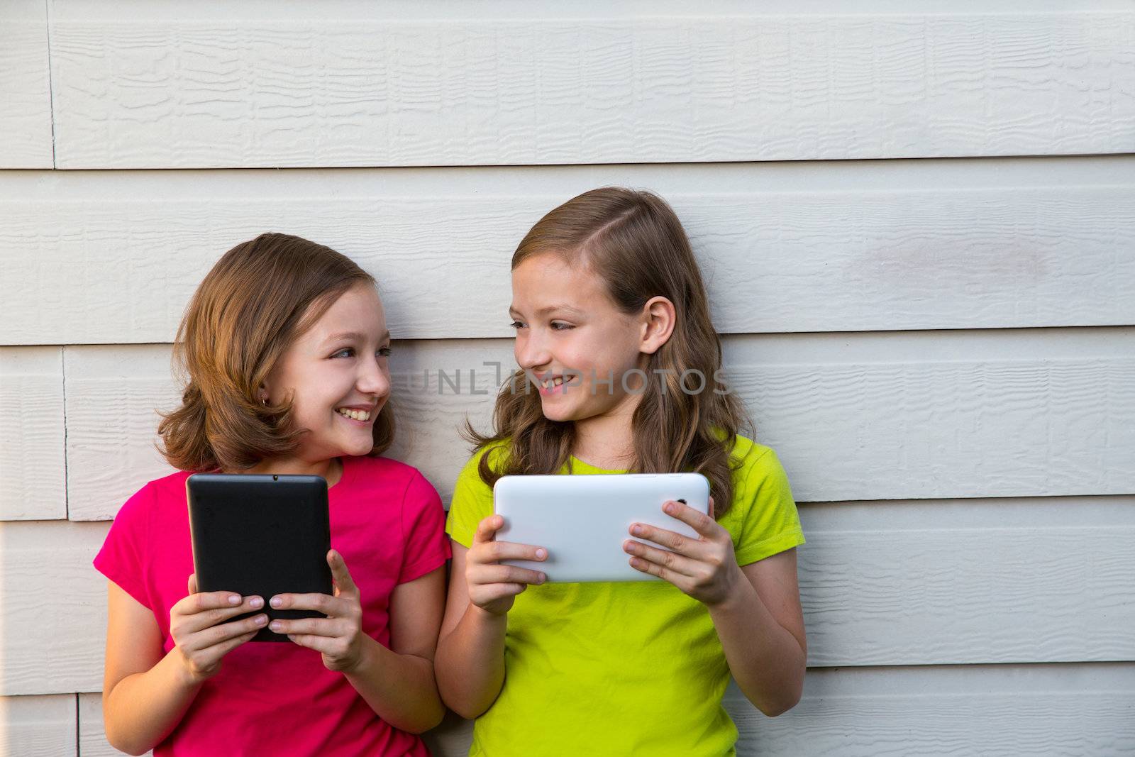 Twin sister girls playing with tablet pc happy on white wall looking each other