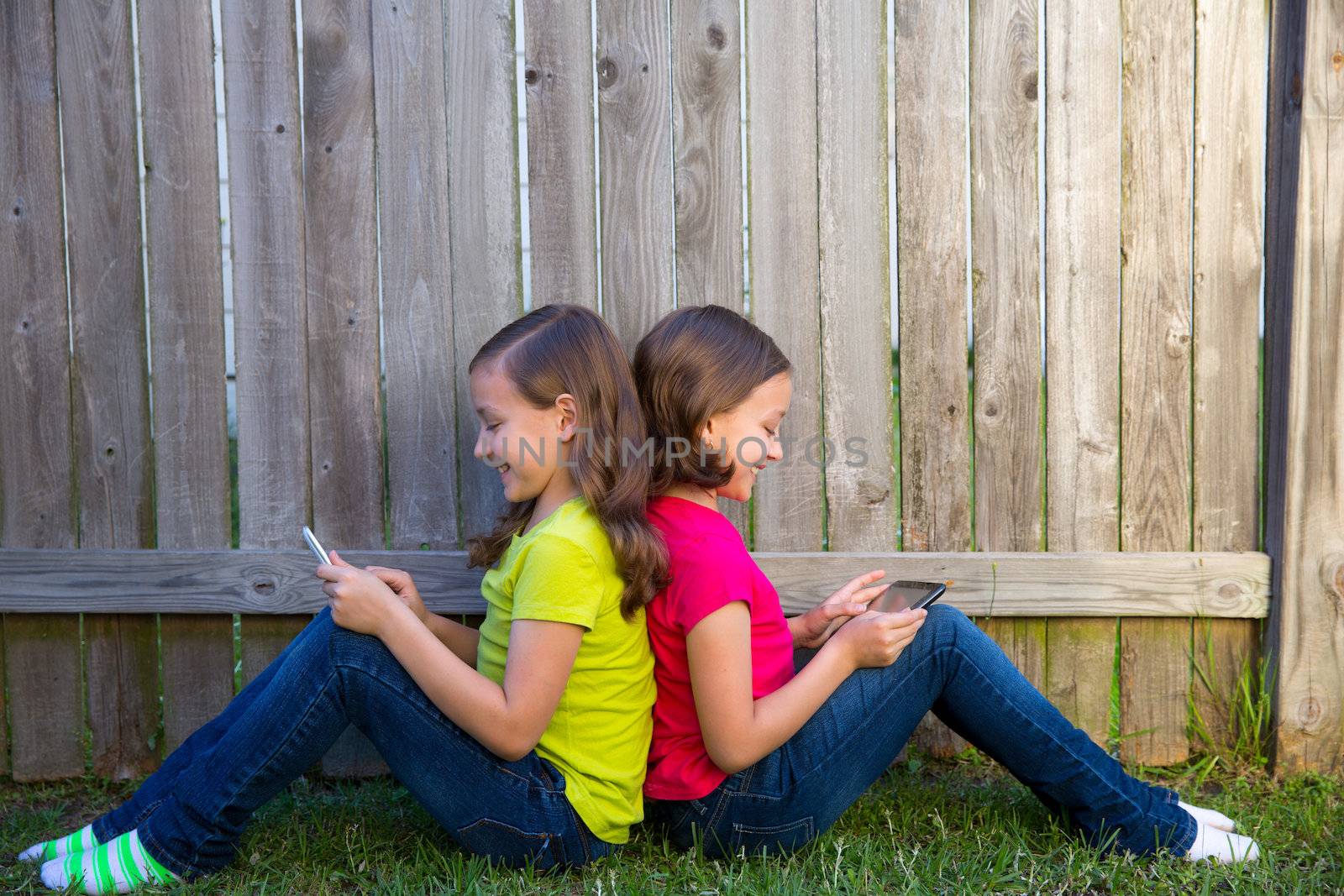 Twin sister girls playing with tablet pc sitting on backyard lawn fence leaning on her back