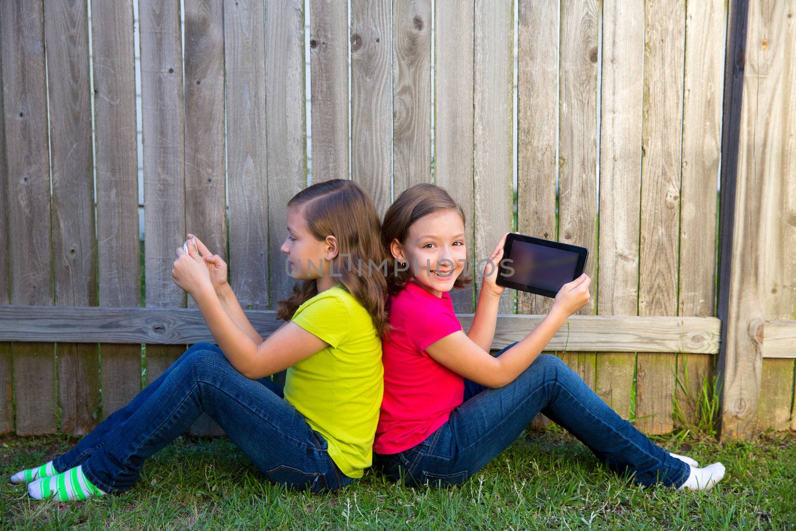 Twin sister girls playing with tablet pc sitting on backyard lawn fence leaning on her back
