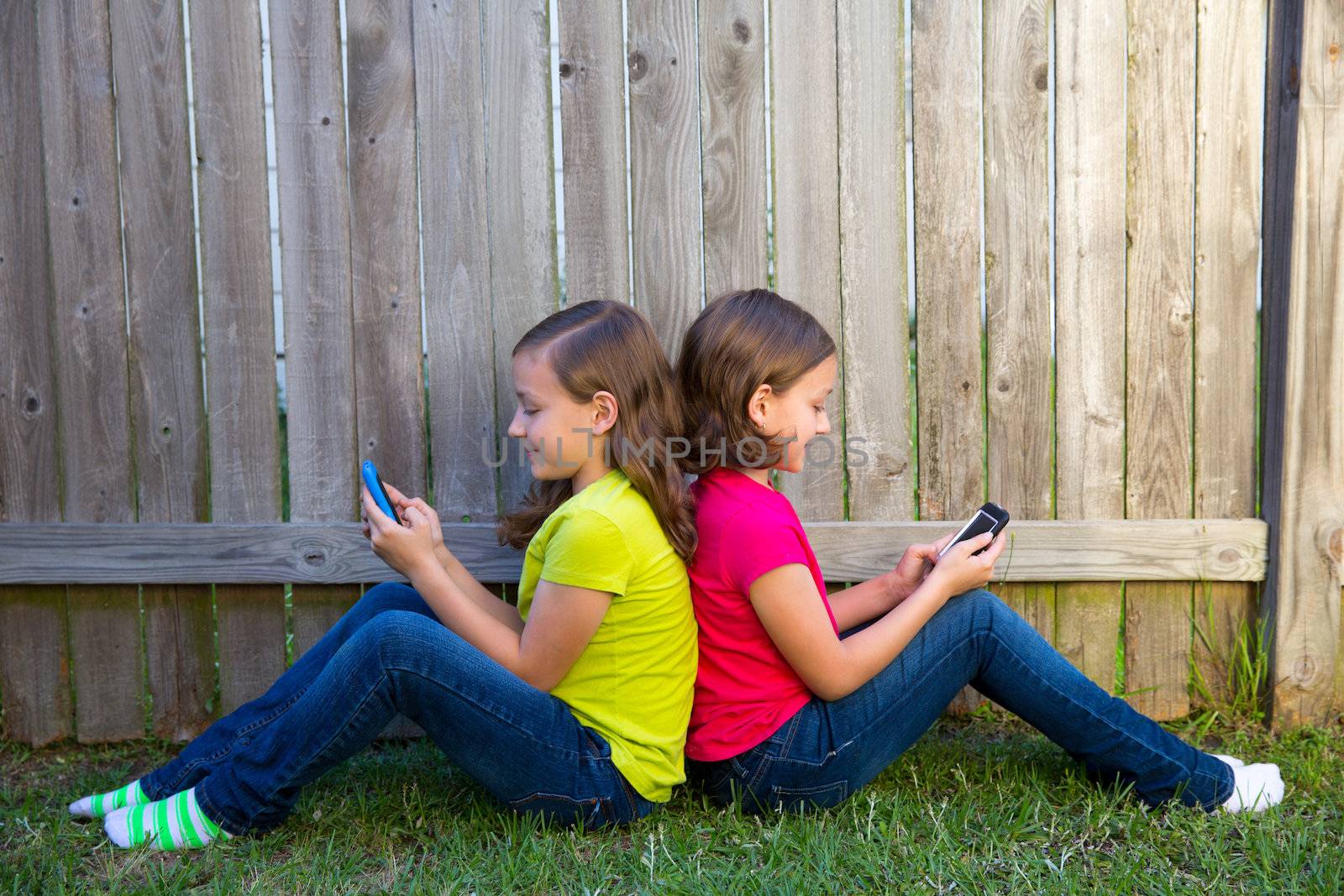 Twin sister girls playing smartphone sitting on backyard lawn by lunamarina