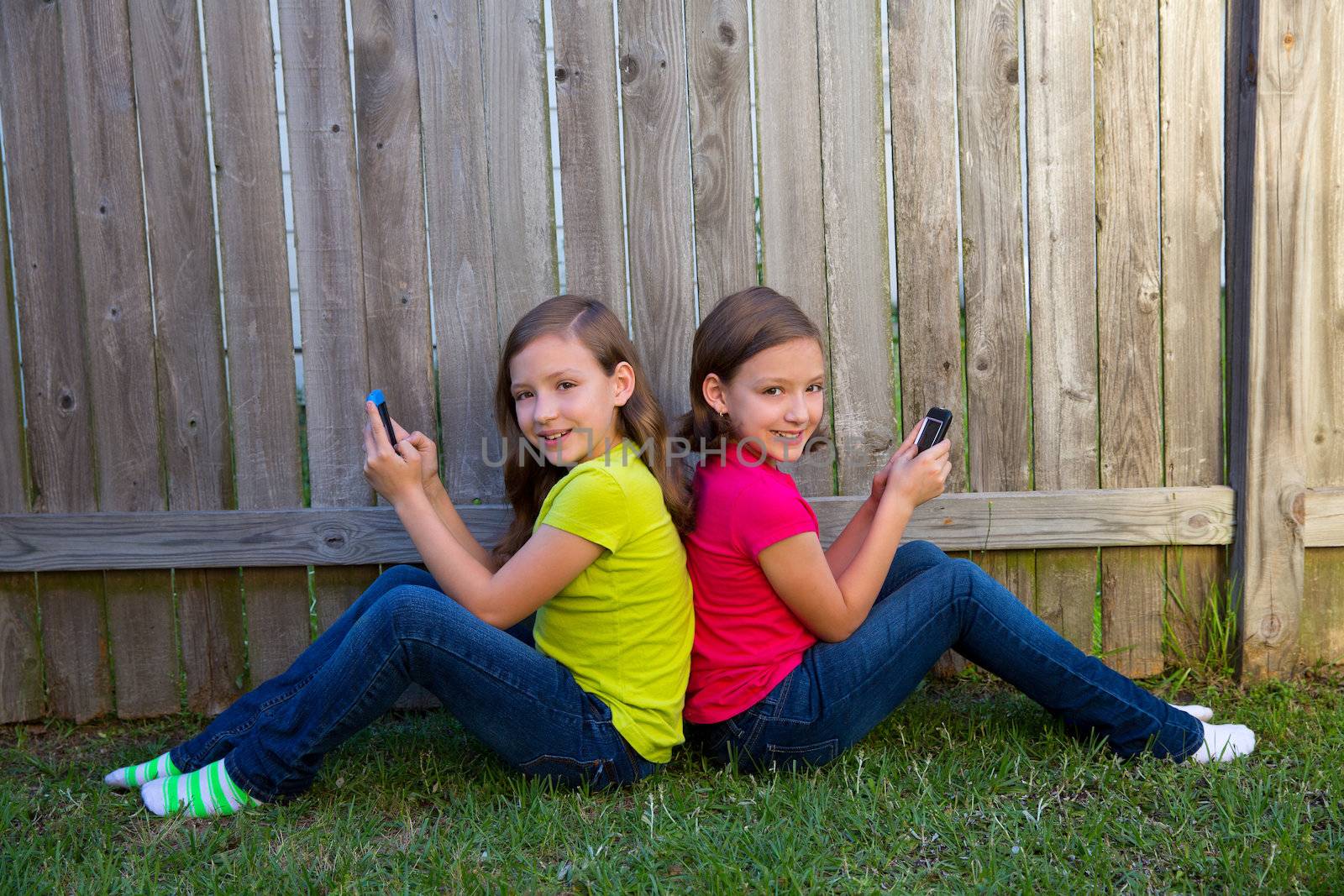 Twin sister girls playing with smartphone sitting on backyard lawn fence leaning on her back