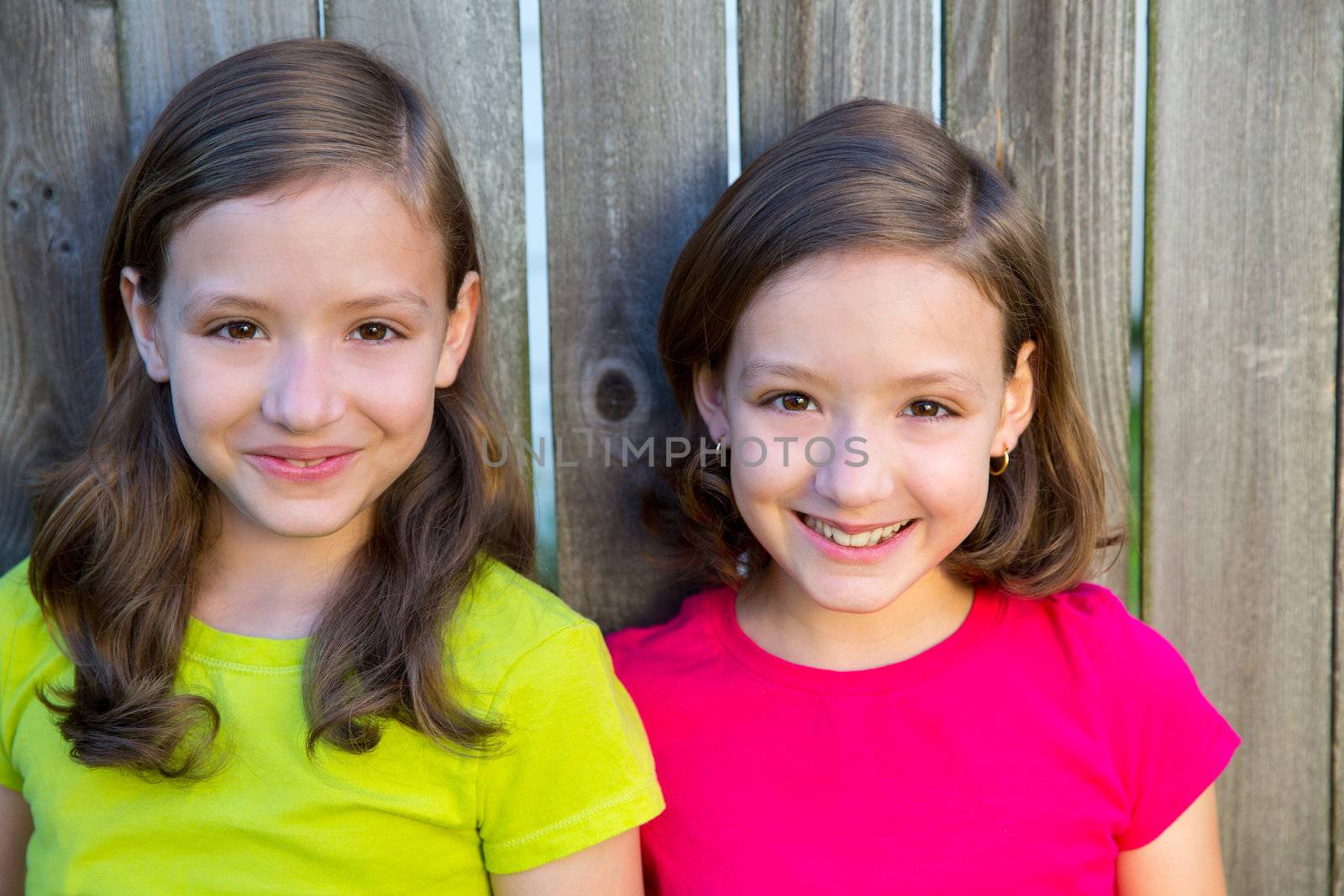 Happy twin sisters with different hairstyle smiling on wood backyard fence