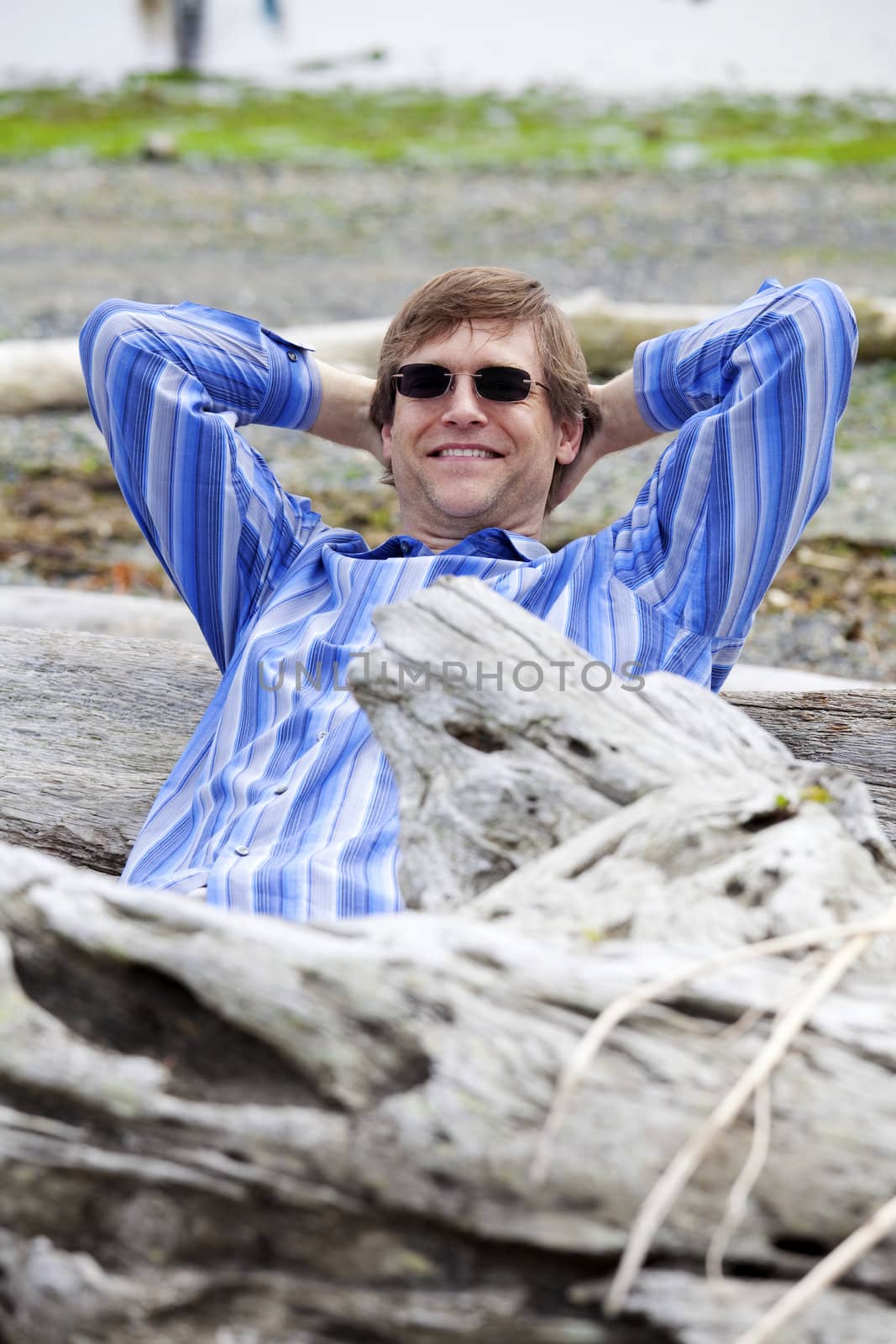 Handsome Caucasian man in forties leaning back against log on beach, relaxed with arms behind head.