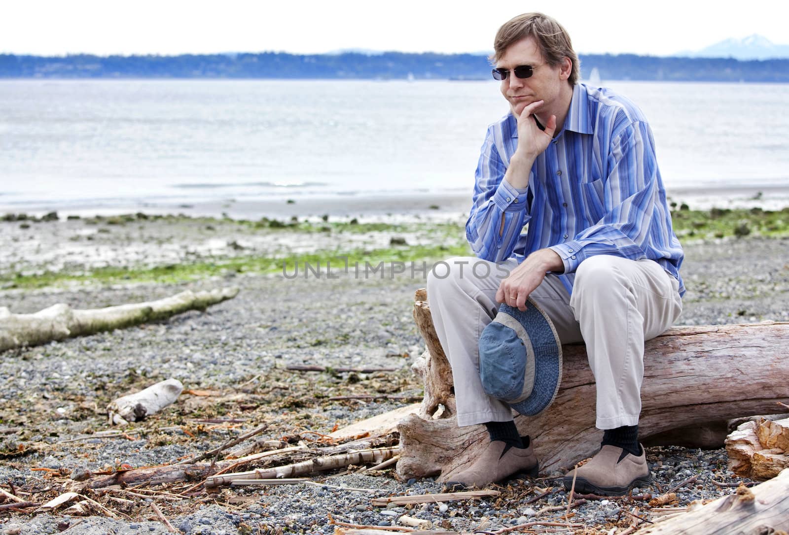 Depressed, sad Caucasian  man in forties sitting on driftwood on beach, chin in hand