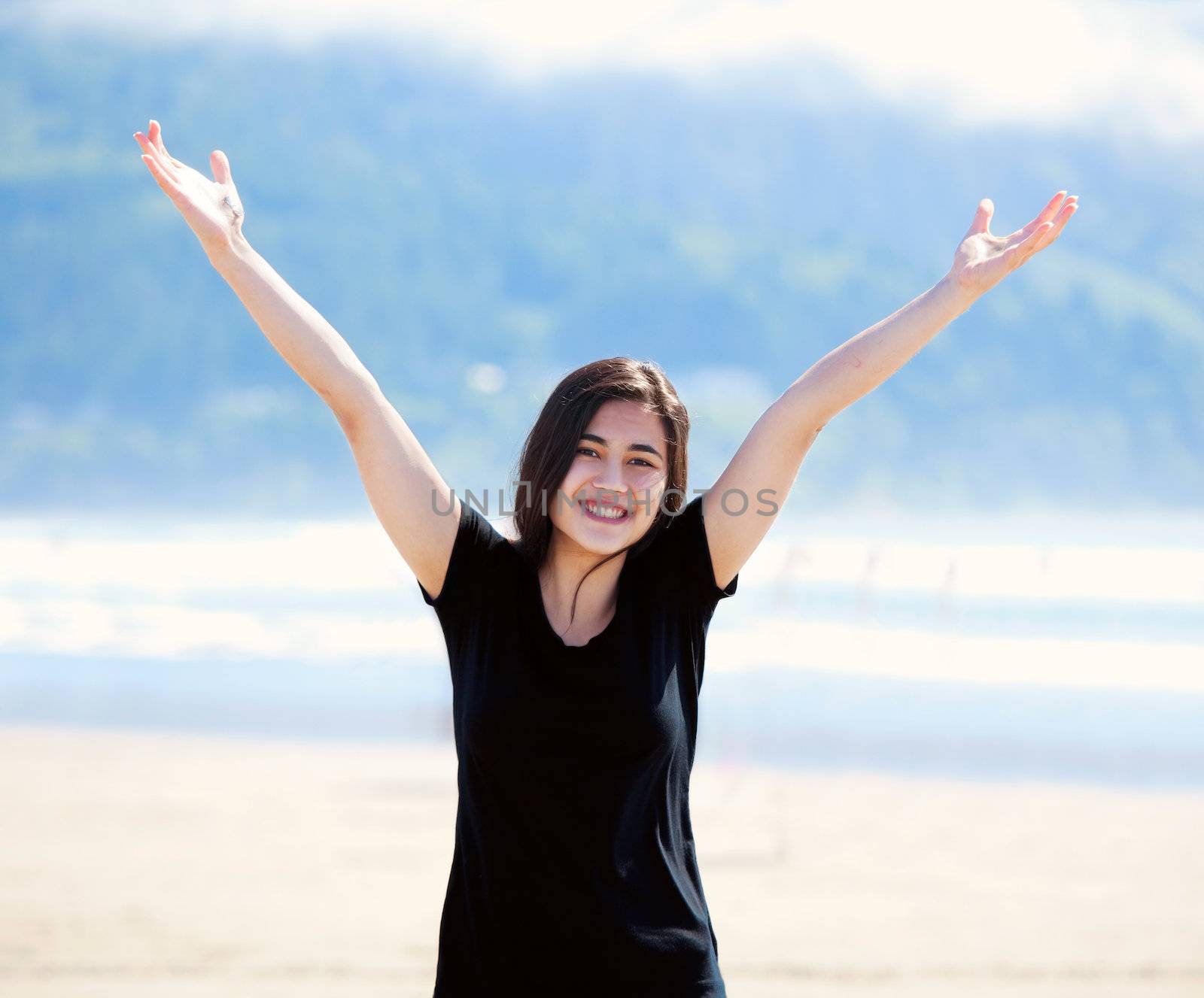 Happy young biracial woman on beach, arms outstretched to the sky, smiling