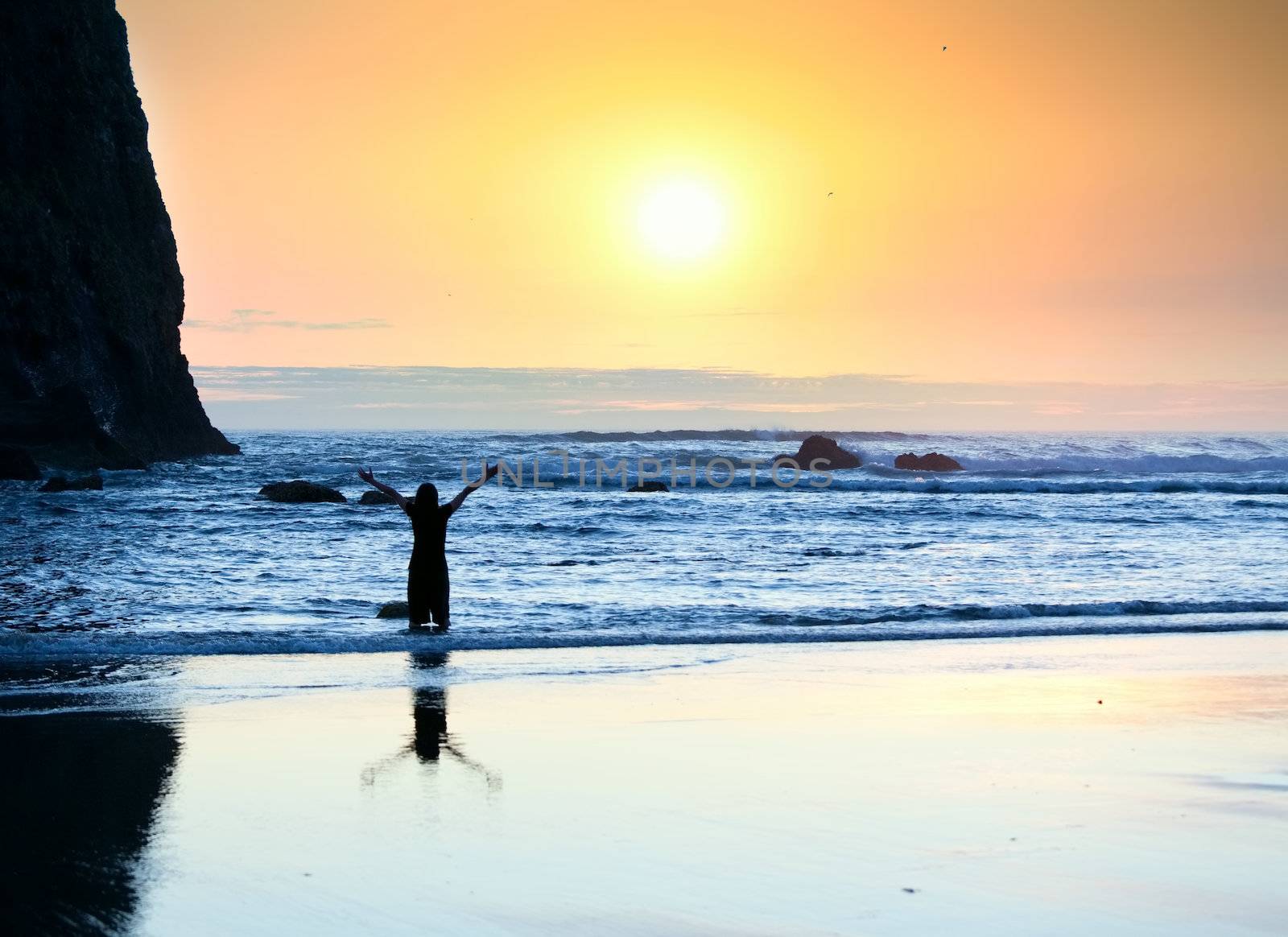 Silhouette of girl standing in waves, arms raised in praise to God at sunset
