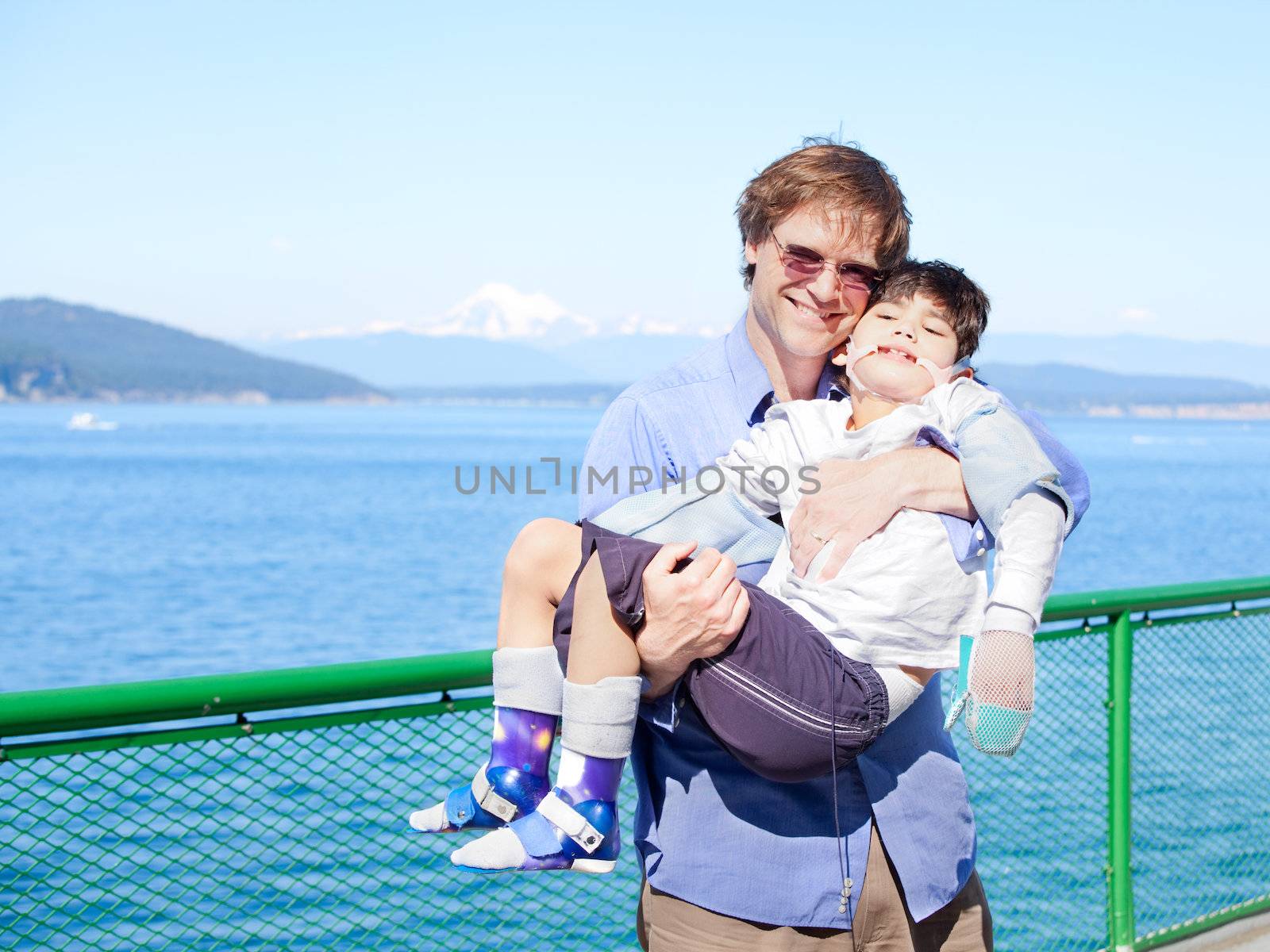 Father holding disabled son in arms on deck of ferry boat. Puget Sound in background. Child has cerebral palsy.