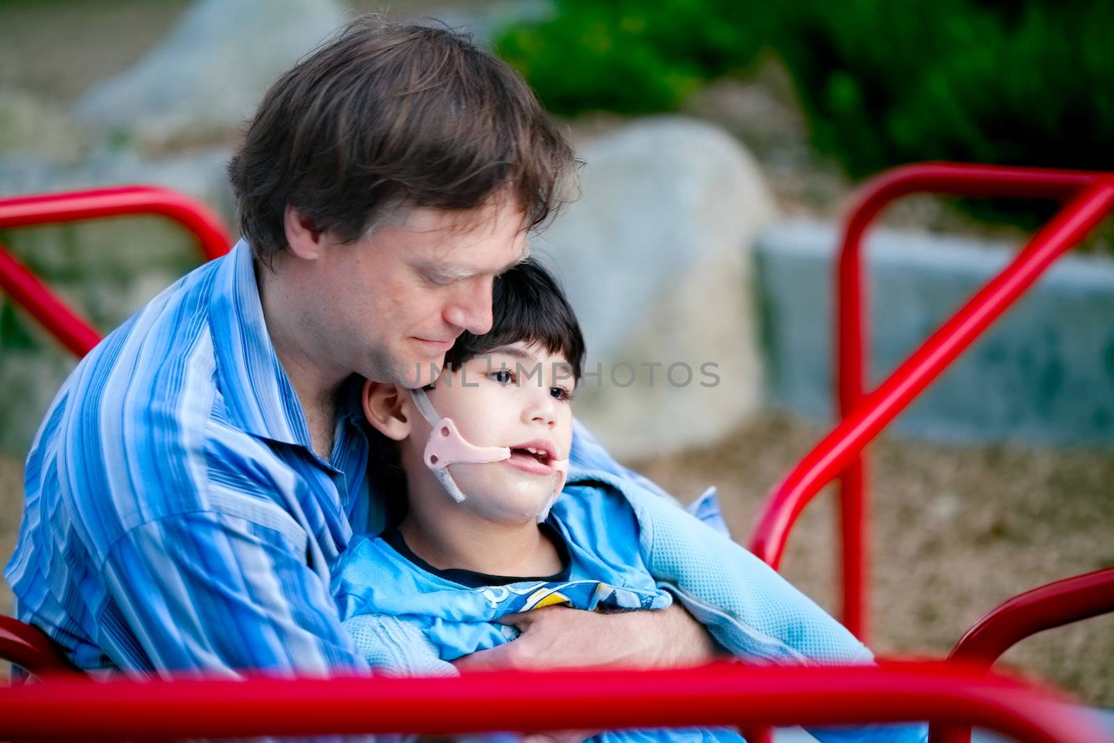 Father playing with disabled son on merry go round at playground. Child has cerebral palsy. 
