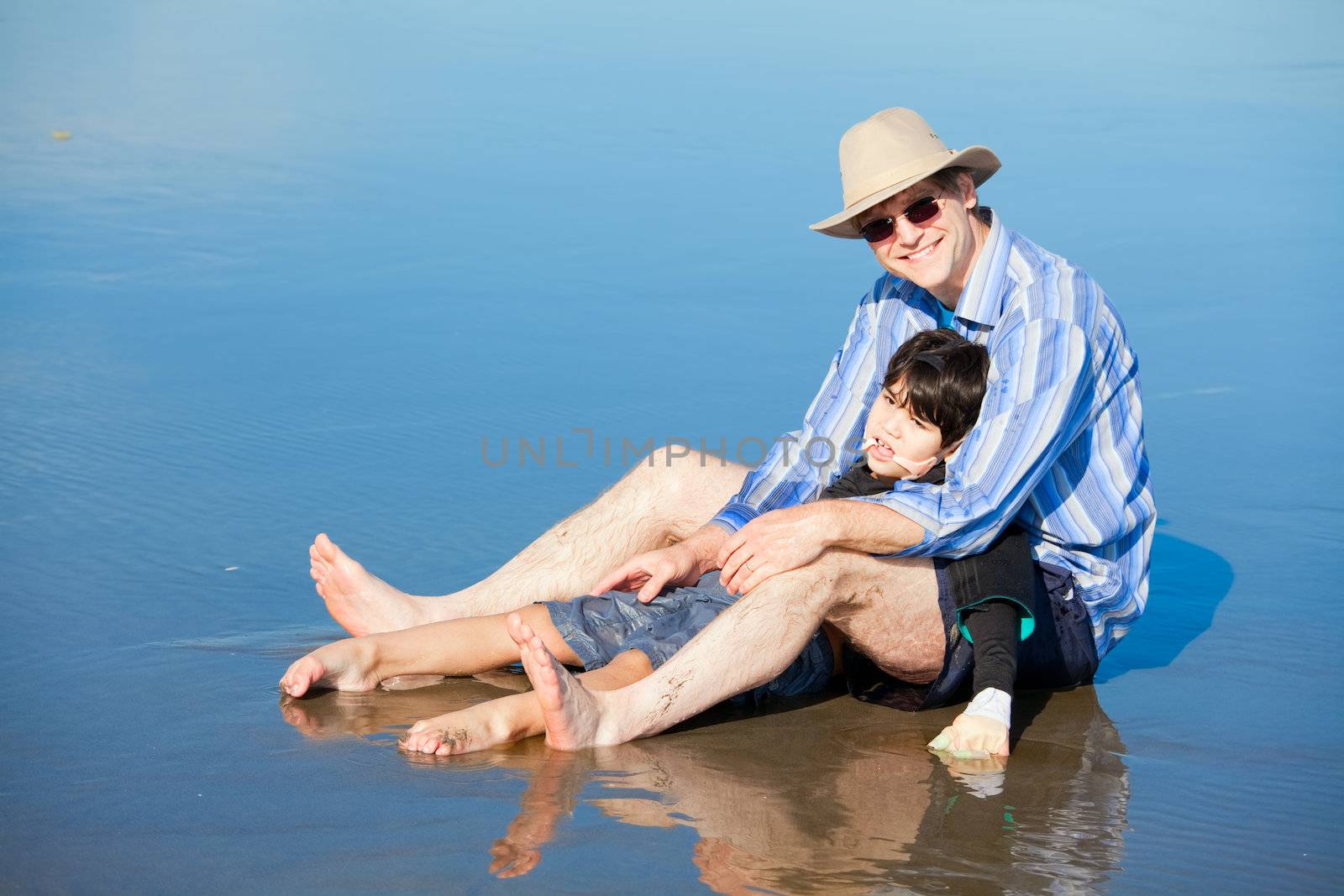 Father playing with disabled son on beach, holding him upright. Child has cerebral palsy