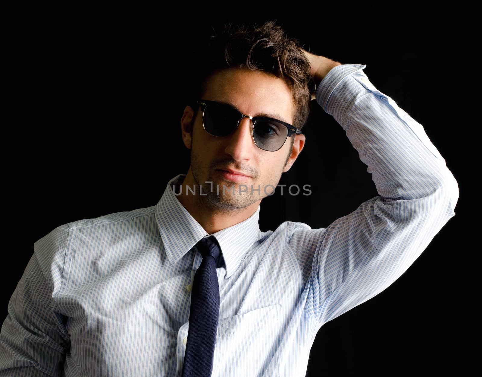 Handsome, elegant young man with shirt, tie and sunglasses, one hand on his head, isolated on black