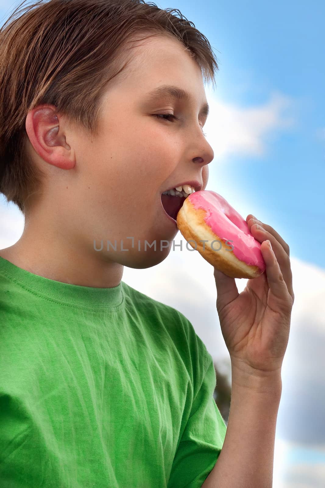 A hungry young child biting a pink strawberry iced doughnut.   He is wearing a green t-shirt.  Sky and clouds background