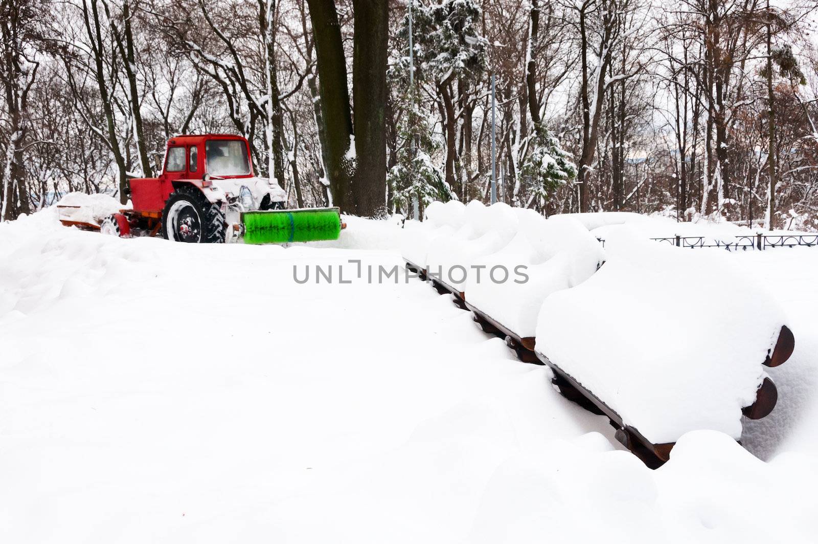 Red tractor cleaning winter park after snowfall with wooden benches on front