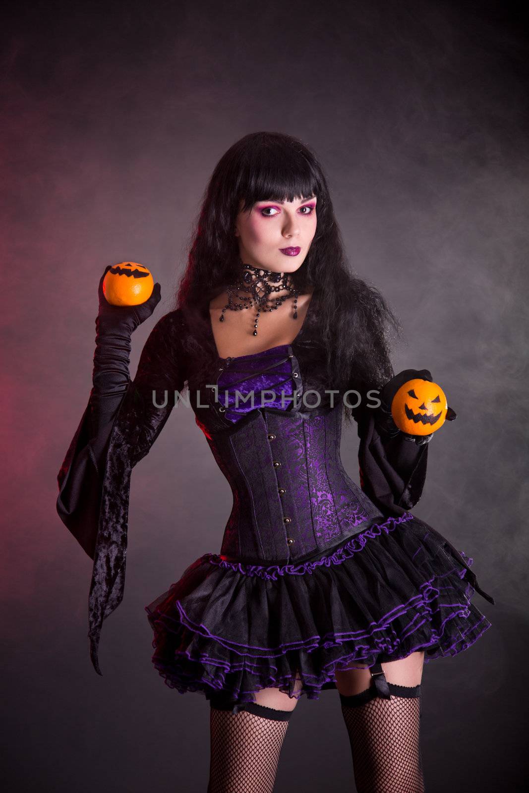 Smiling witch in purple gothic Halloween costume holding Jack-o-lantern style oranges 