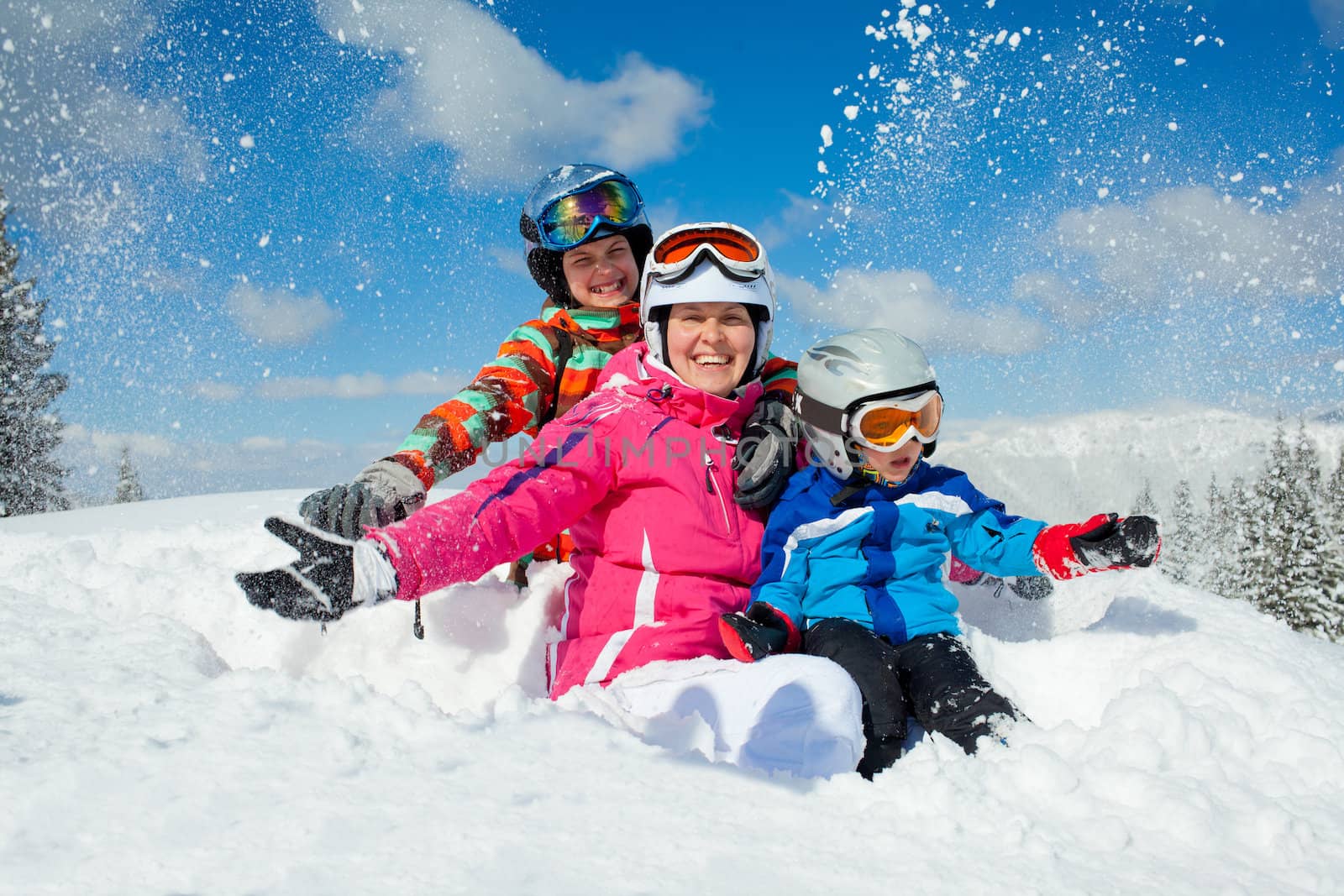 Skiing, winter, family - smiling boy in ski goggles and a helmet with his mother and sister playing in snow in winter resort