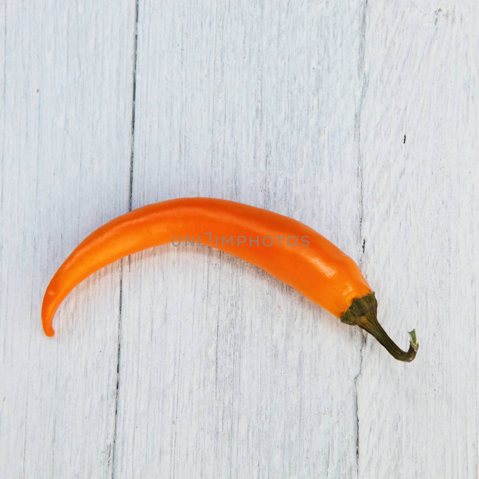 Single whole fresh crescent-shaped orange capsicum pepper used as a spice, flavouring and vegetable in cooking on a white painted rustic wooden plank