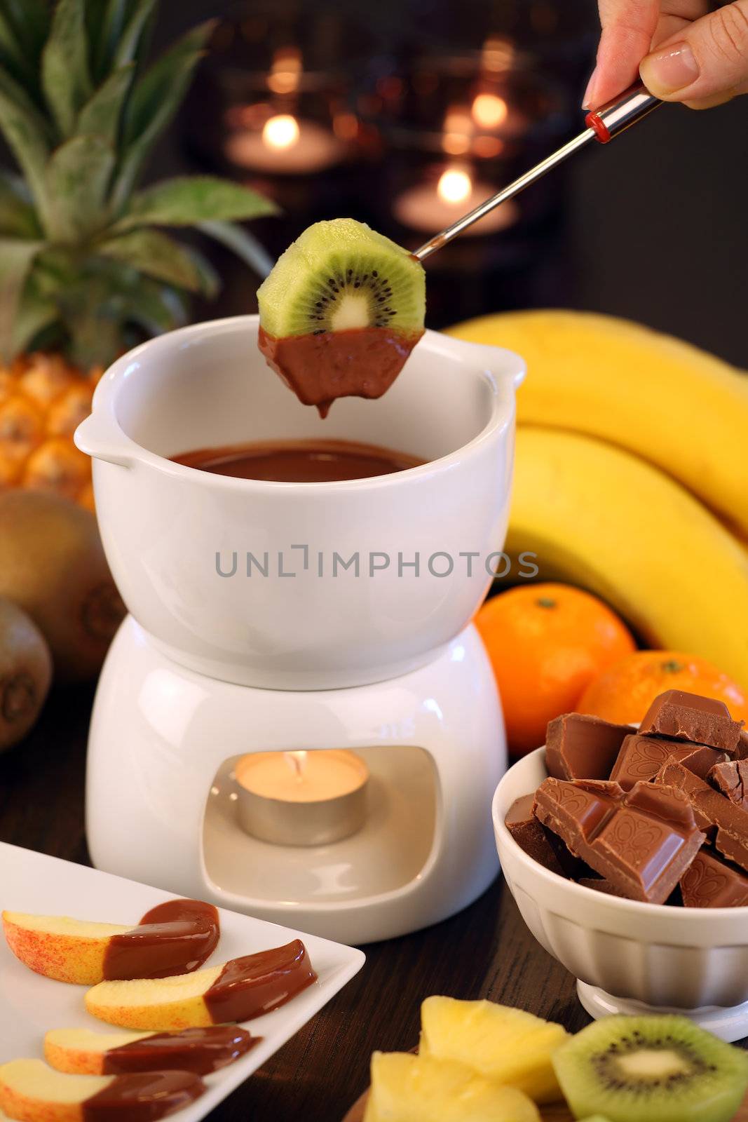 Photo of a kiwi slice being dipped into the bowl of the chocolate fondue. Selective focus on the kiwi.