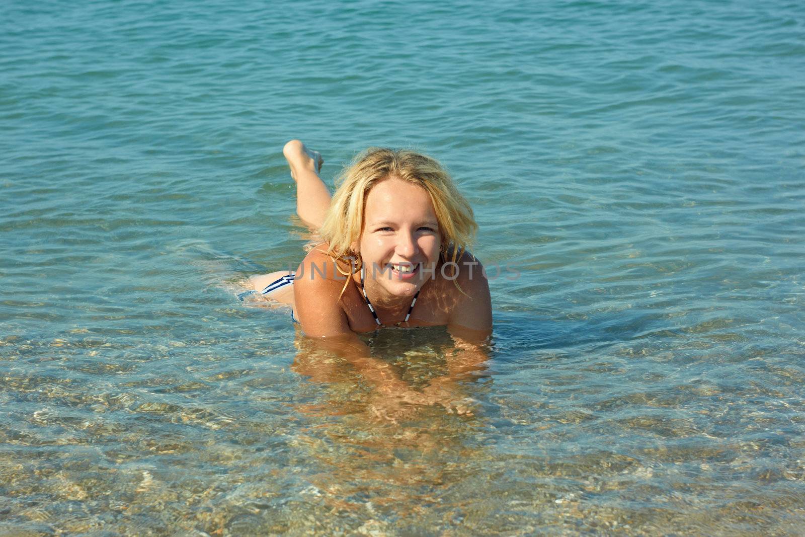 Beautiful smiling teenage girl lying in the shallow coastal sea water with pebbly bottom in sunny weather