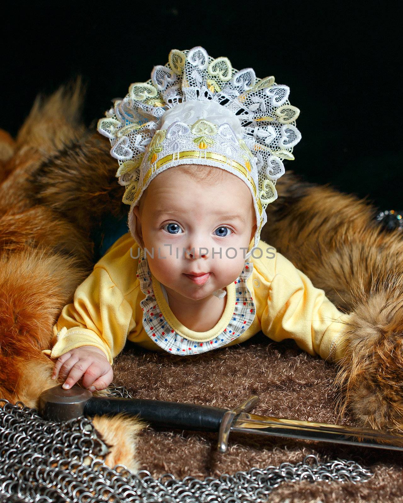 Blue-eyed baby lying on fur litter near the hauberk and sword, fox pelt in the background