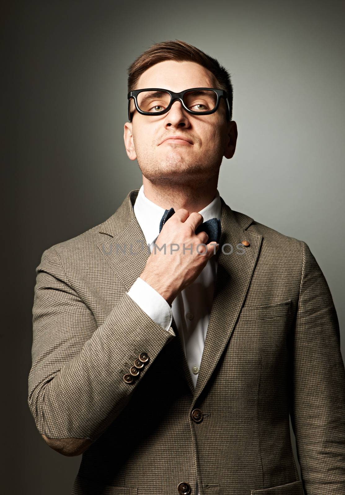 Confident nerd in eyeglasses adjusting his bow-tie against grey background