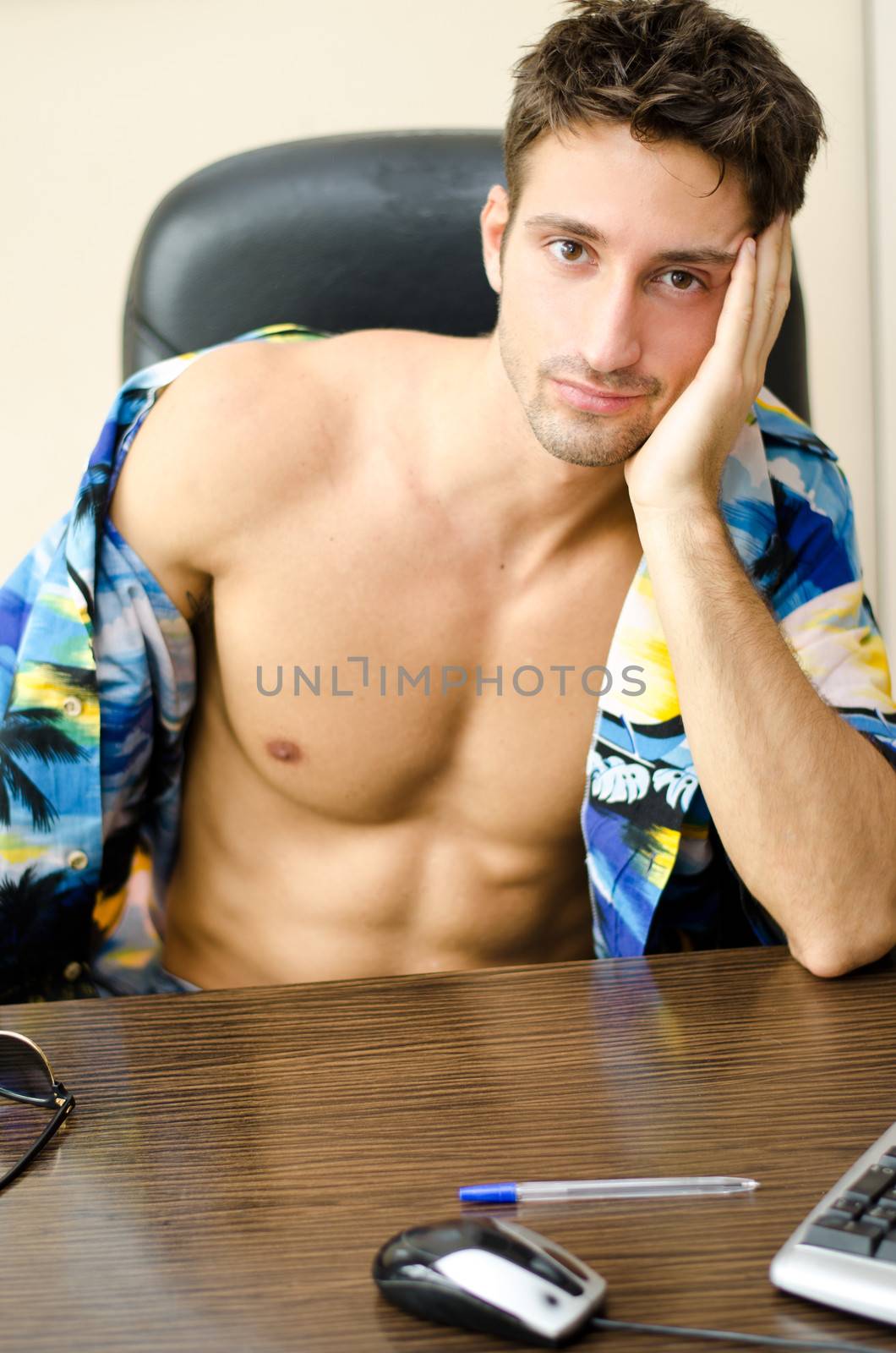 Attractive, fit young man with his shirt open sitting at office desk barechested thinking about vacation