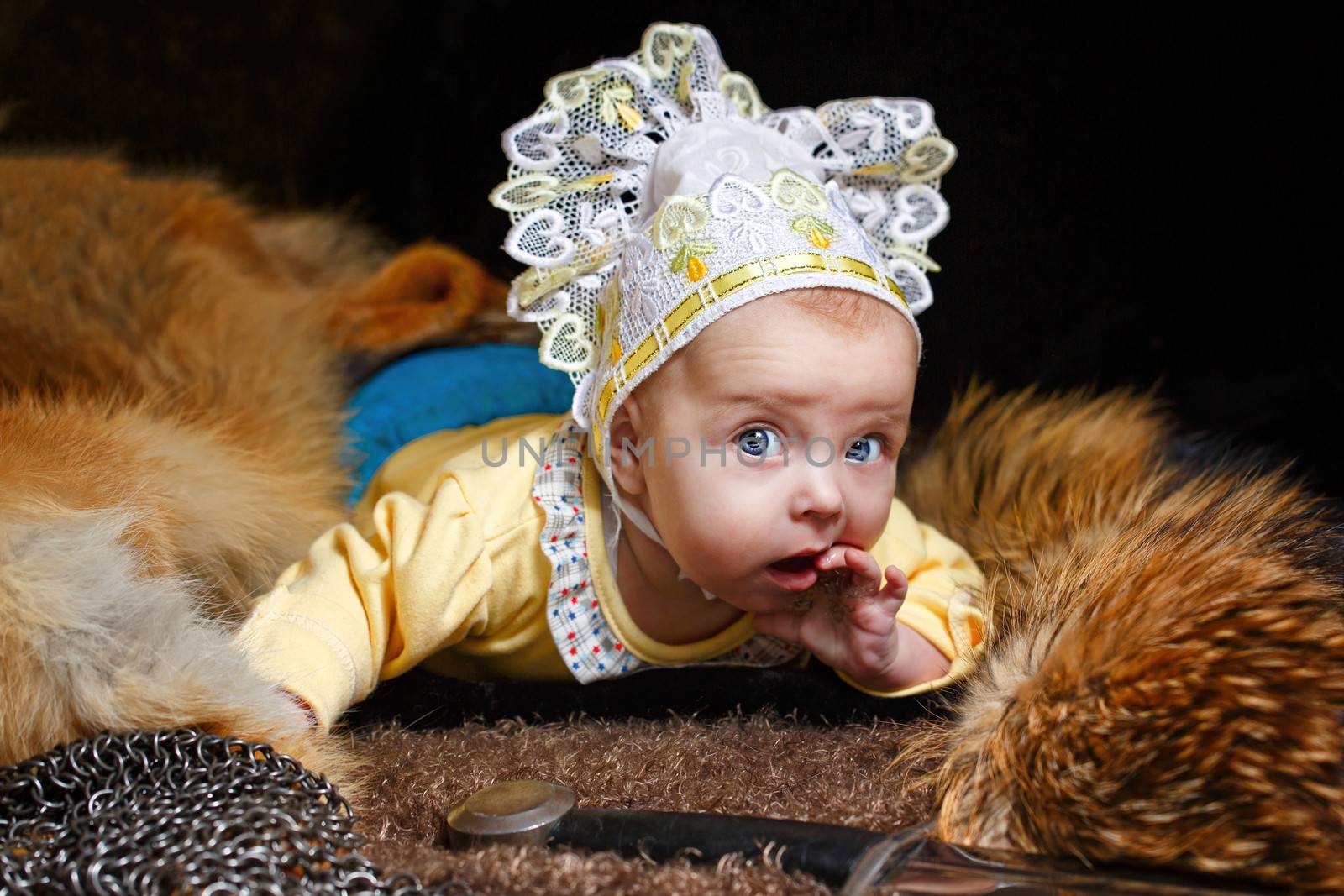 Blue-eyed baby lying on fur litter near the hauberk and sword, fox pelt in the background