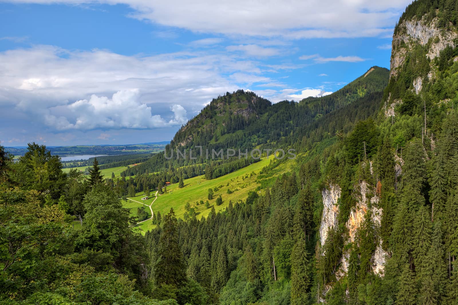 View of sunlit green meadow surrounded by forest-covered mountains under beautiful cloudy sky in Bavaria, Germany.