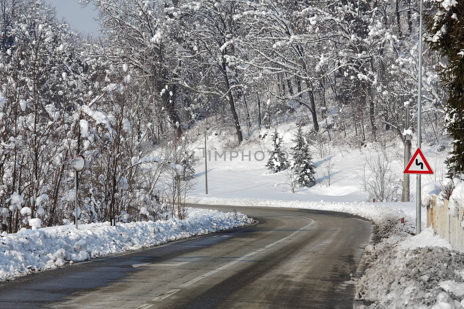 Gray curved road through trees and hills covered by snow at winter in Piedmont, Northern Italy.