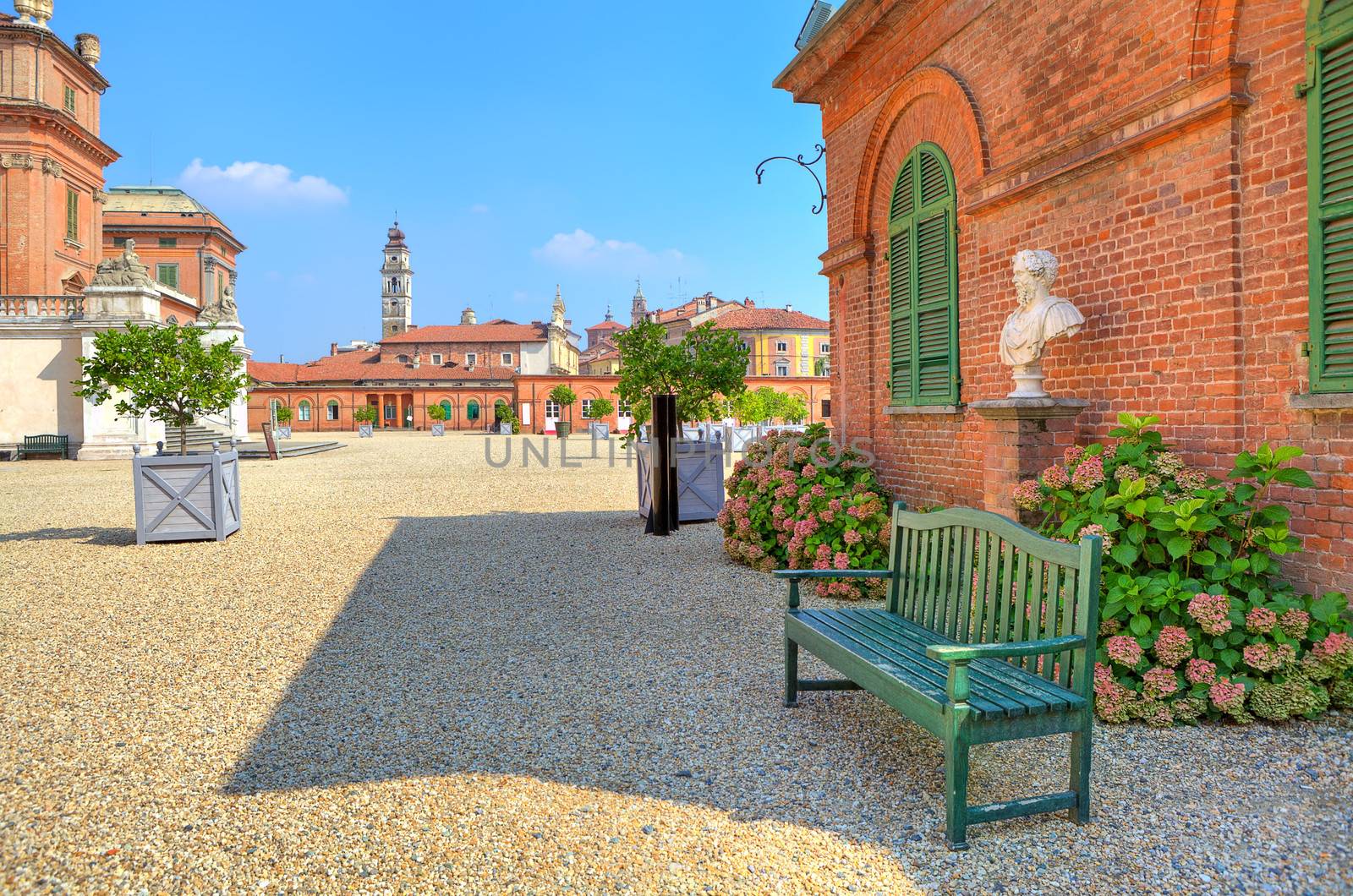 Wooden bench on the gravel pathway next to red brick house at the entrance to park and Royal Castle of Racconigi in Piedmont, Northern Italy.